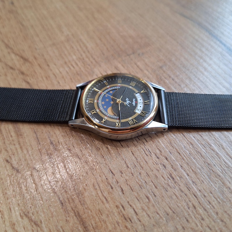 May include: A wristwatch with a black face, gold-colored bezel, and a black mesh band. The watch face features Roman numerals, a moon phase indicator, and the text "Luch". The watch is displayed on a wooden surface.