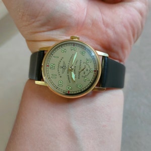 Puede incluir: Un reloj de pulsera dorado con una correa de cuero negro. La esfera del reloj es de color crema con marcas negras y presenta el texto "ШТУРМАНСКИЕ" y "ПЕРВЫЙ ЧЕЛОВЕК В КОСМОСЕ ЮРИЙ ГАГАРИН АПРЕЛЯ 1961".