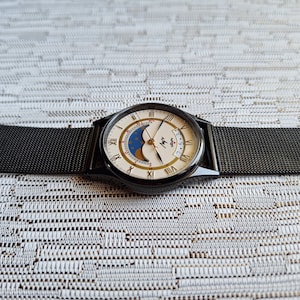May include: A black and white analog wristwatch with a black mesh band. The watch face features a moon phase display and the text "Myy" with a small star below it.