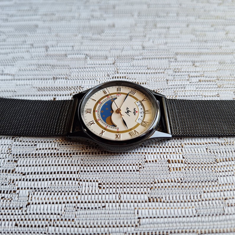 May include: A black and white analog wristwatch with a black mesh band. The watch face features a moon phase display and the text "Myy" with a small star below it.