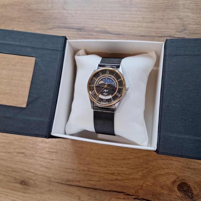 May include: A wristwatch with a black mesh band and a gold-toned bezel. The watch face is black with gold Roman numerals and a moon phase indicator. The watch is presented in a black box with a white pillow.