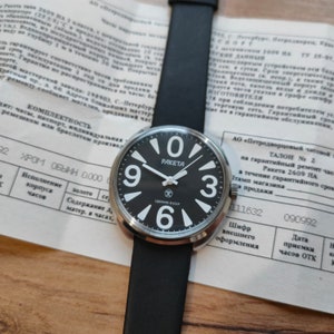 May include: A black Raketa wristwatch with white numerals and hands, displaying the text "PAKETA" and "СДЕЛАНО В СССР" on the face. The watch has a black leather strap and is set against a document background.