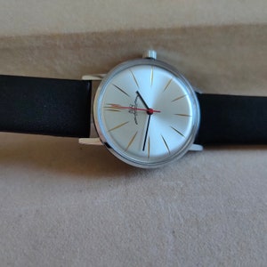 May include: A silver wristwatch with a white face and gold accents. The watch has black hands and a red second hand. The watch is on a black leather band.
