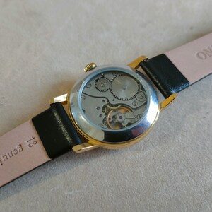 Puede incluir: Un reloj de pulsera dorado con una correa de cuero negro. La esfera del reloj es transparente y muestra el funcionamiento interno del reloj.