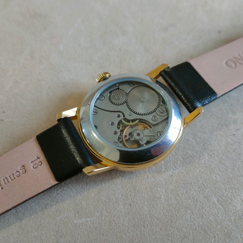 Puede incluir: Un reloj de pulsera dorado con una correa de cuero negro. La esfera del reloj es transparente y muestra el funcionamiento interno del reloj.