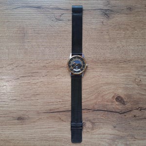 May include: A wristwatch with a black mesh band and a gold-colored bezel. The watch face features a dark background with a moon phase design and the brand name. The watch is displayed on a wooden surface.
