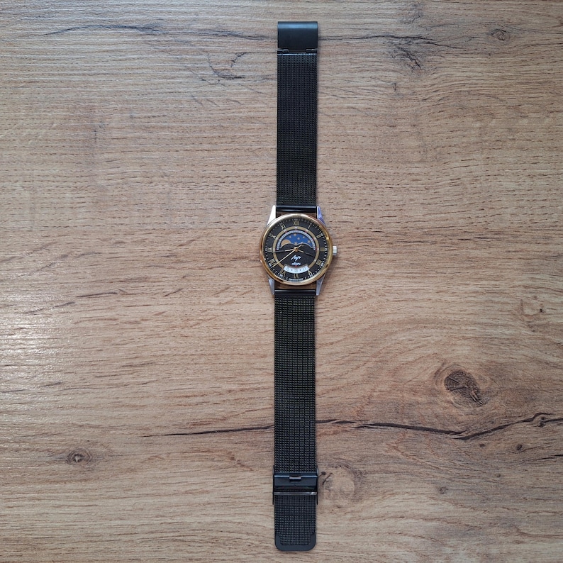 May include: A wristwatch with a black mesh band and a gold-colored bezel. The watch face features a dark background with a moon phase design and the brand name. The watch is displayed on a wooden surface.