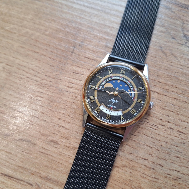 May include: A vintage wristwatch with a black mesh band and a round face. The watch face features gold-tone accents, Roman numerals, and a moon phase design with a blue and gold color scheme. The watch is set against a wooden surface.
