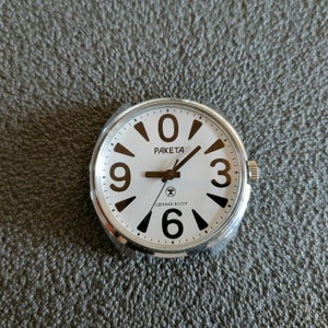 May include: A vintage Raketa watch face with a white dial and black numerals. The watch face has a silver-coloured metal casing and black hands. The text "RAKETA" and "СДЕЛАНО В СССР" are printed on the dial.
