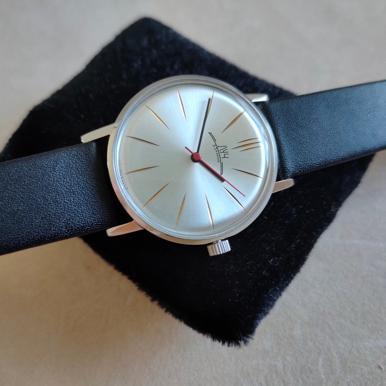 May include: A silver wristwatch with a white face and black leather band. The watch face has gold-colored hour markers and a red second hand. The watch face also has the text "HWU 23 Jewels" printed on it.