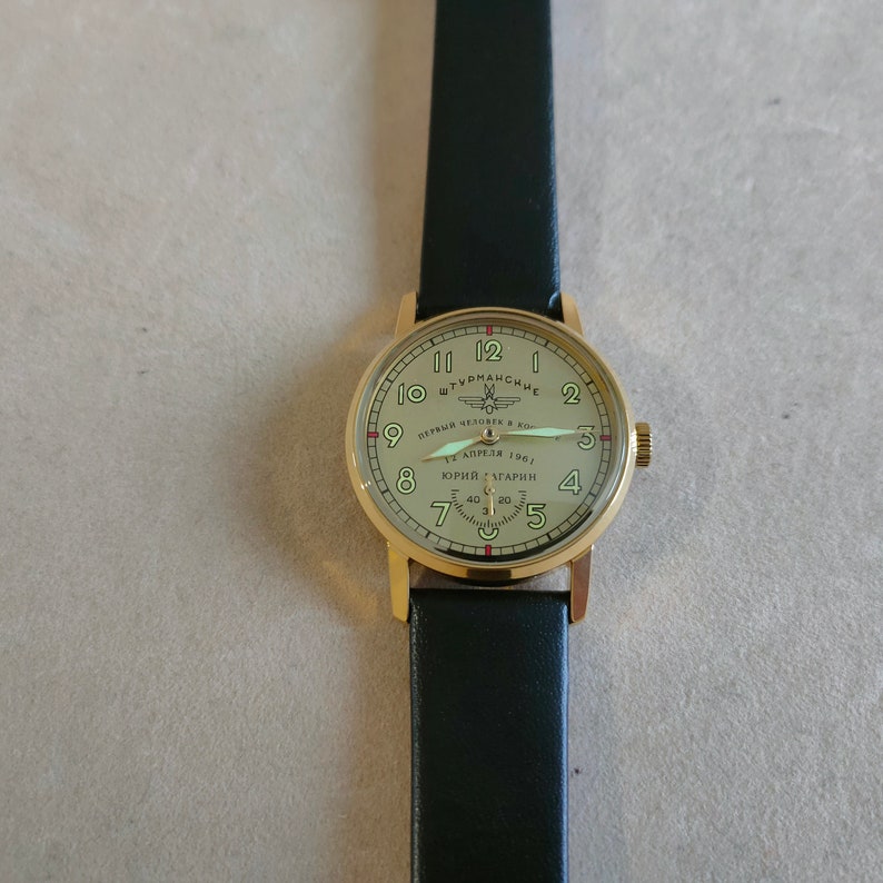 Puede incluir: Un reloj de pulsera dorado con una correa de cuero negro. La esfera del reloj es blanca con marcas negras y presenta el texto "ШТУРМАНСКИЕ" y "ПЕРВЫЙ ЧЕЛОВЕК В КОСМОСЕ" con una estrella roja y un peque&ntilde;o gr&aacute;fico de avi&oacute;n. El texto "12 АПРЕЛЯ 1961" y "ЮРИЙ ГАГАРИН" tambi&eacute;n est&aacute; en la esfera del reloj.