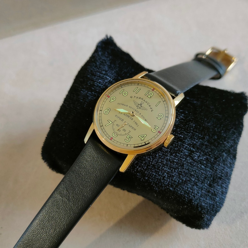 Puede incluir: Un reloj de pulsera dorado con una correa de cuero negra. La esfera del reloj es blanca con n&uacute;meros negros y una aguja de segundos roja. La esfera del reloj tambi&eacute;n presenta el texto "12 апреля 1961", "ЮРИЙ ГАГАРИН", y "СССР".