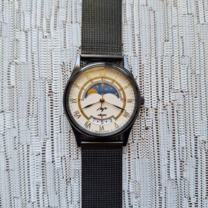 May include: A black wristwatch with a silver face and gold accents. The watch has a moon phase complication and a date window. The watch is on a black mesh band. The watch face says "часы" and "Сделано в СССР".