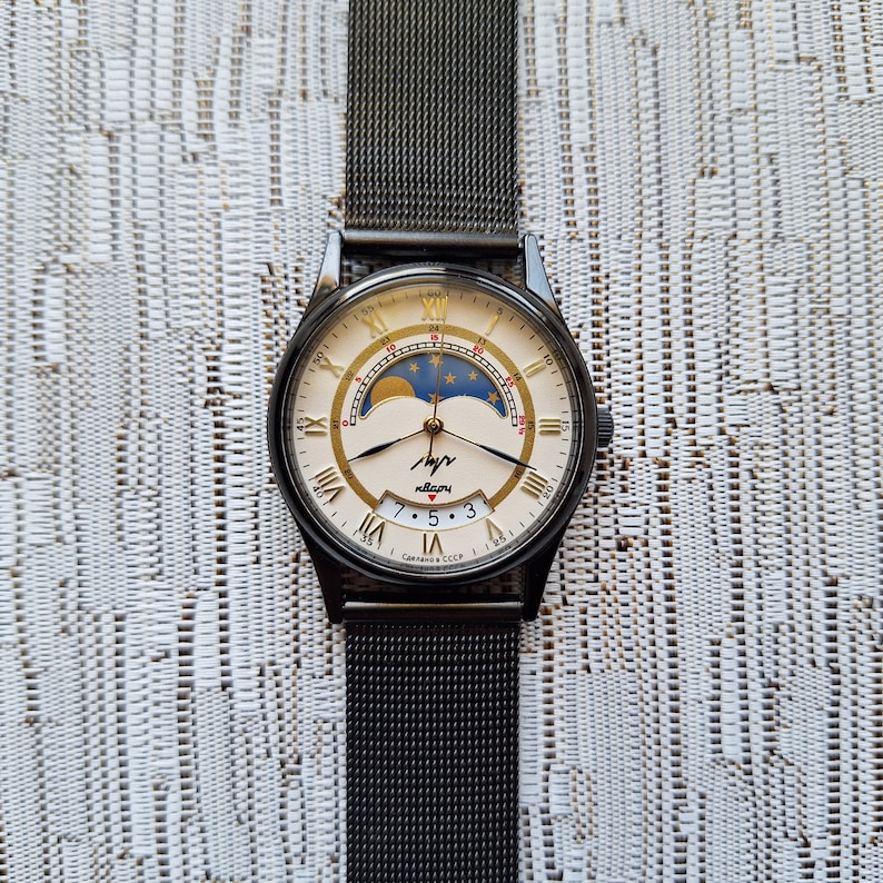 May include: A black wristwatch with a silver face and gold accents. The watch has a moon phase complication and a date window. The watch is on a black mesh band. The watch face says "часы" and "Сделано в СССР".
