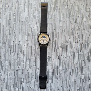May include: A black watch with a silver face and a black mesh band. The watch face has a moon phase display and a date window.