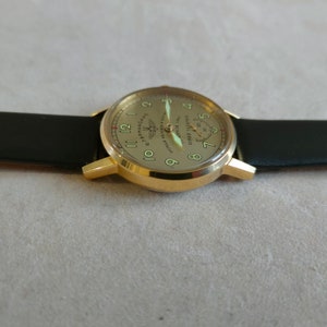 Puede incluir: Un reloj de pulsera dorado con una correa de cuero negro. La esfera del reloj es de color crema con n&uacute;meros negros y un peque&ntilde;o subdial. La esfera del reloj tambi&eacute;n presenta el texto "АПРЕЛЯ 1941 ПЕРВЫЙ ЧЕЛОВЕК В КОСМОСЕ ЮРИЙ ГАГАРИН" en escritura cir&iacute;lica.