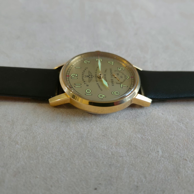 Puede incluir: Un reloj de pulsera dorado con una correa de cuero negro. La esfera del reloj es de color crema con n&uacute;meros negros y un peque&ntilde;o subdial. La esfera del reloj tambi&eacute;n presenta el texto "АПРЕЛЯ 1941 ПЕРВЫЙ ЧЕЛОВЕК В КОСМОСЕ ЮРИЙ ГАГАРИН" en escritura cir&iacute;lica.