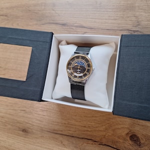 May include: A wristwatch with a black mesh band and a gold-toned bezel. The watch face is dark with gold Roman numerals and a moon phase indicator. The watch is presented in a black box with a white pillow.