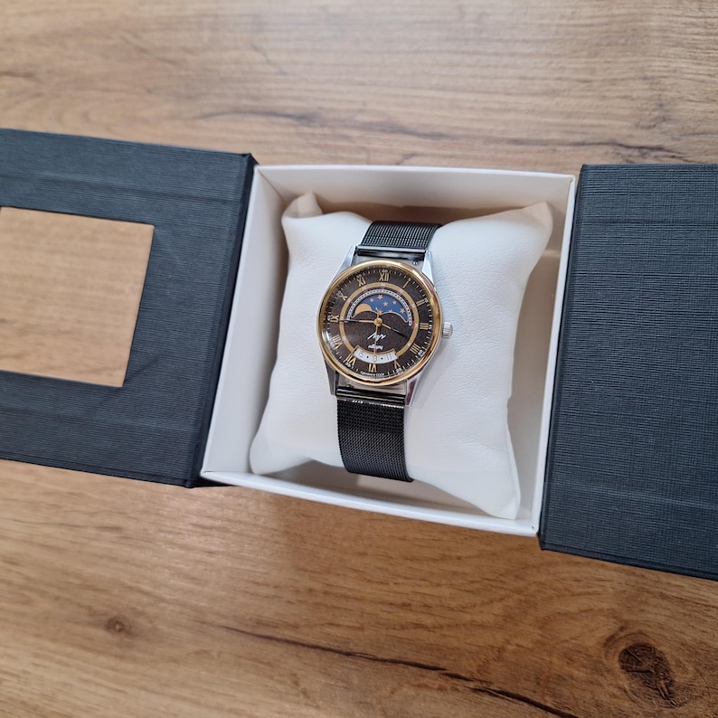 May include: A wristwatch with a black mesh band and a gold-toned bezel. The watch face is dark with gold Roman numerals and a moon phase indicator. The watch is presented in a black box with a white pillow.