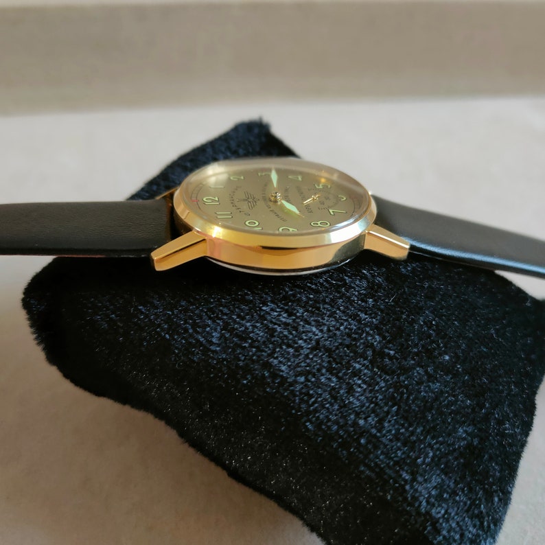 Puede incluir: Un reloj de pulsera dorado con una correa de cuero negro. La esfera del reloj tiene un fondo blanco con marcadores de hora en n&uacute;meros romanos negros y una aguja de segundos verde.