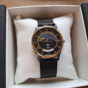 May include: A vintage wristwatch with a black face, gold-tone bezel, and a black mesh band. The watch face features Roman numerals, a moon phase indicator, and the text "Лучь". The watch is presented in a white box.