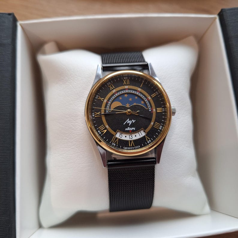May include: A vintage wristwatch with a black face, gold-tone bezel, and a black mesh band. The watch face features Roman numerals, a moon phase indicator, and the text "Лучь". The watch is presented in a white box.
