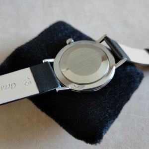 May include: A silver wristwatch with a black leather band. The watch is on a black velvet cushion. The back of the watch is visible, showing a silver case with a brushed finish.