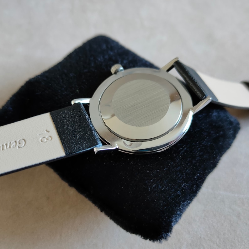 May include: A silver wristwatch with a black leather band. The watch is on a black velvet cushion. The back of the watch is visible, showing a silver case with a brushed finish.