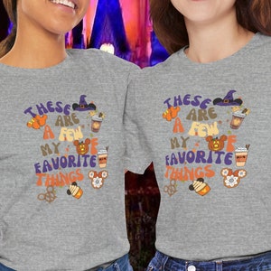 Pode incluir: Duas pessoas vestindo camisetas cinzas com o texto "These are a few of my favorite things" e vários ícones temáticos de outono. As camisetas são de cor cinza mesclada e têm um visual desgastado. As pessoas estão em pé em frente a um prédio iluminado à noite.