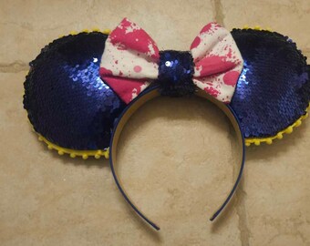 Op de afbeelding: Blauwe en gele pailletten Minnie Mouse oren met een roze en witte strik met witte spetters.