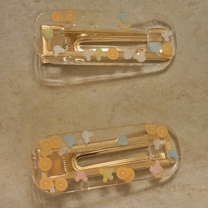 May include: Two clear plastic hair clips with gold metal clasps. The clips are decorated with small, colourful, confetti-like shapes.