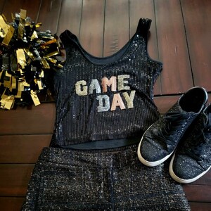 May include: Black sequined tank top with the words "GAME DAY" in gold, pink, and white. The tank top is paired with a black sequined skirt.