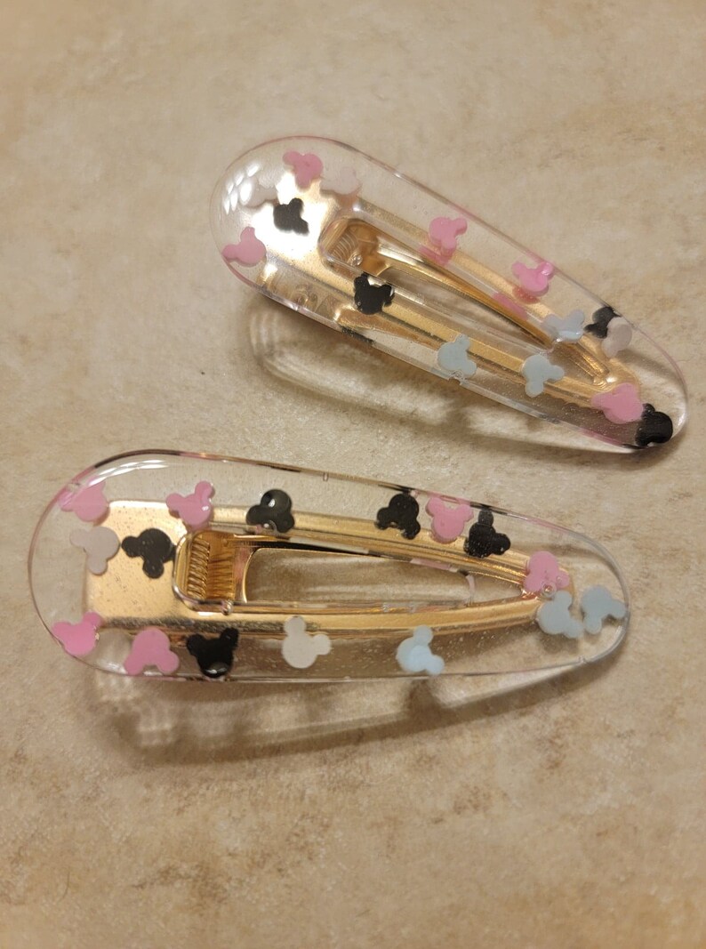May include: Two clear plastic hair clips with gold metal clasps. The clips are decorated with small, colourful Mickey Mouse heads.