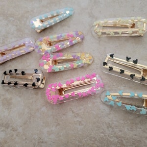 May include: A set of ten clear plastic hair clips with different coloured confetti and Mickey Mouse designs. The clips are all the same size and shape, and they have a gold metal spring.