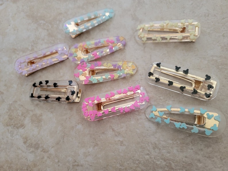 May include: A set of ten clear plastic hair clips with different coloured confetti and Mickey Mouse designs. The clips are all the same size and shape, and they have a gold metal spring.