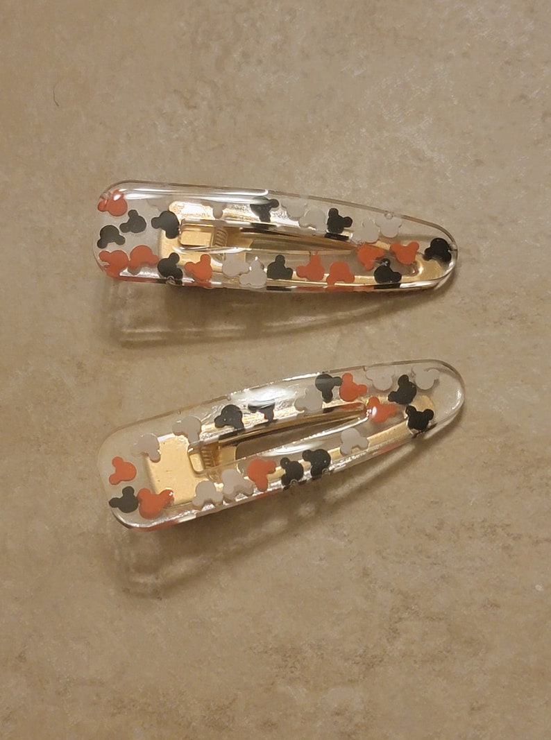 May include: Two clear plastic hair clips with a gold metal spring. The clips are decorated with small, colourful Mickey Mouse heads.