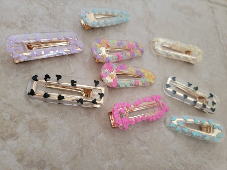 May include: A set of ten clear plastic hair clips with different coloured designs. The designs include pink, yellow, blue, and black Mickey Mouse heads.