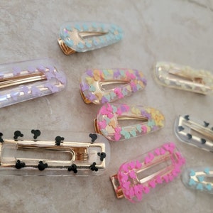 May include: A set of nine clear plastic hair clips with different coloured glitter and designs. The clips have a gold metal spring and are approximately 5 cm long.