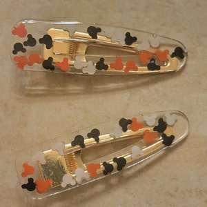 May include: Two clear plastic hair clips with gold metal clasps. The clips are decorated with small, colourful Mickey Mouse heads in black, white, and orange.
