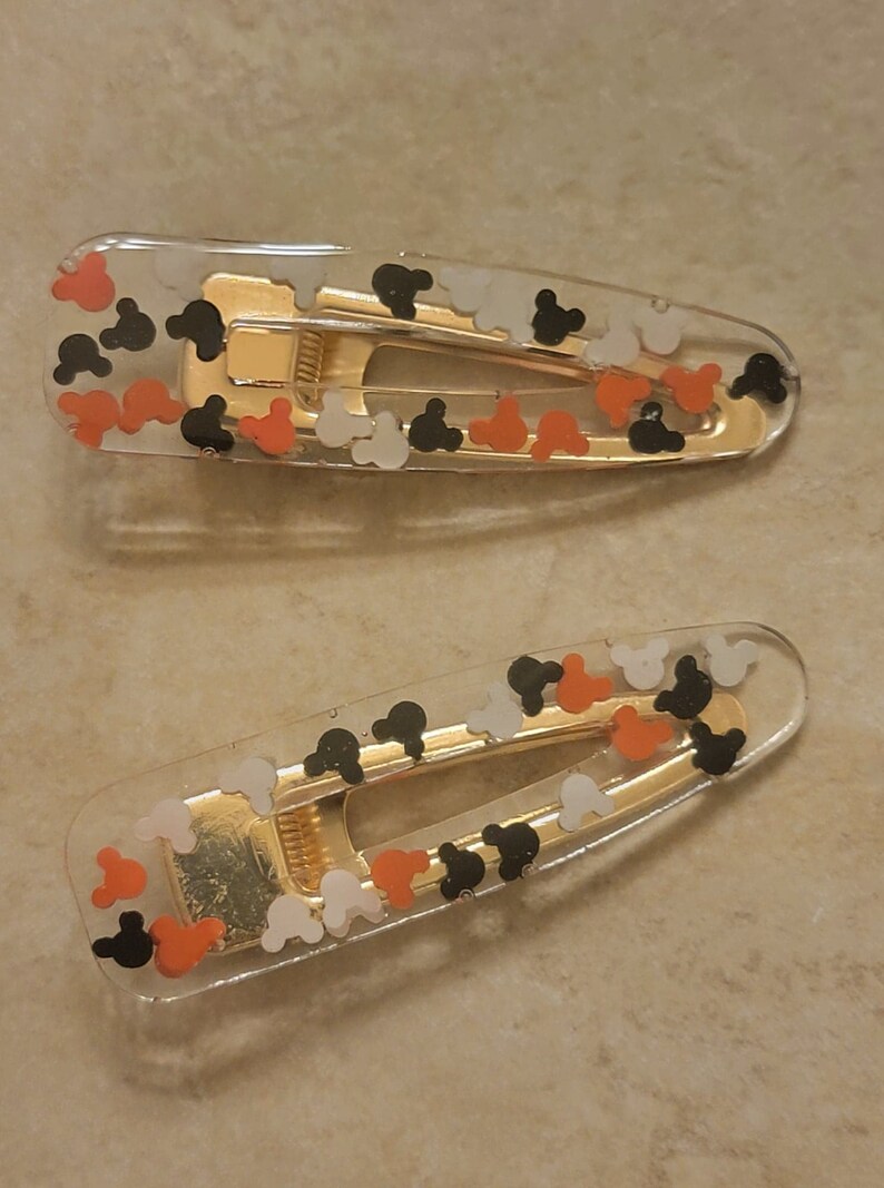 May include: Two clear plastic hair clips with gold metal clasps. The clips are decorated with small, colourful Mickey Mouse heads in black, white, and orange.