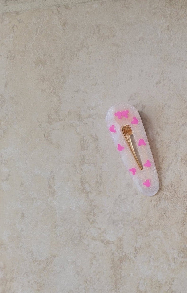 May include: A white hair clip with a gold metal bar and pink Mickey Mouse silhouettes.