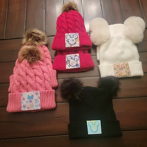 May include: Four knit hats with pom-poms and various Disney character patches. The hats are pink, red, white, and black.