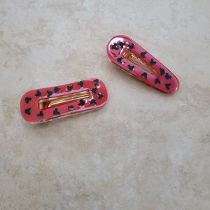 May include: Two red hair clips with black Mickey Mouse silhouettes. The clips are oval shaped and have a gold metal spring.