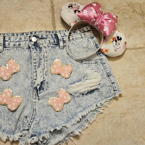 May include: Light blue denim shorts with pink bow appliques. The shorts have a frayed hem and distressed details.
