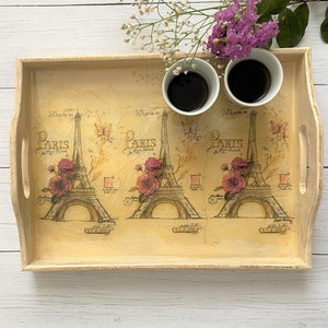 May include: A wooden tray with a Paris theme featuring three Eiffel Tower illustrations and floral accents. The tray is painted in a light beige color and has two handles. Two coffee cups are on the tray.