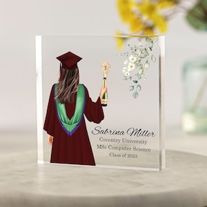 Può includere: Un blocco di vetro trasparente con un'immagine stampata di una laureata che tiene una bottiglia di champagne. La laureata indossa una toga e un cappello marroni con accenti verdi e viola. Il testo sul blocco recita "Sabrina Miller, Coventry University, MSc Computer Science, Class of 2023".