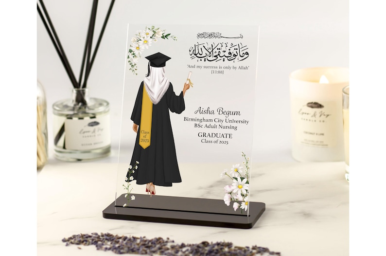 May include: A clear acrylic graduation plaque with a black-gowned graduate and a yellow sash. The plaque displays the graduate's name, university, degree, and graduation year. Floral decorations and Arabic script are also included.