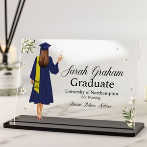 Puede incluir: Una placa de acrílico transparente con una base negra, que presenta a una graduada con una toga y un birrete azules, sosteniendo un diploma. El texto dice "Sarah Graham Graduate University of Northampton BSc Nursing Dream Believe Achieve".
