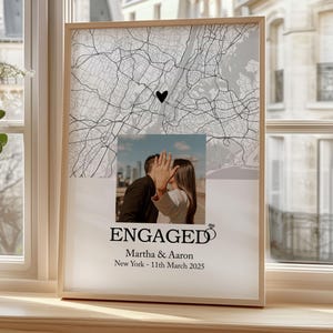May include: A framed engagement announcement. It features a map of New York with a heart marking a location, a photo of a couple, the word "ENGAGED", names, and the date March 11, 2025. The frame is light-coloured.
