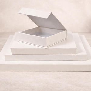 May include: A stack of four square white boxes of varying sizes, arranged with the largest at the bottom and the smallest at the top. The top box is open, revealing a hollow interior, while the three boxes beneath it are closed and flat.